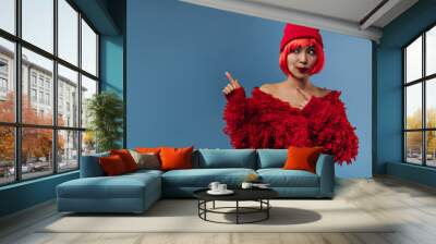 Funny stylish girl with red hair and bright makeup in modern cap and long sleeve cool clothes looking away and showing to place for text.. Wall mural