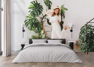 Funny young lady in trendy pants standing on one leg and showing peace sign with smile. Laughing curly girl in stylish sneakers dancing beside big green flower and looking down. Wall mural