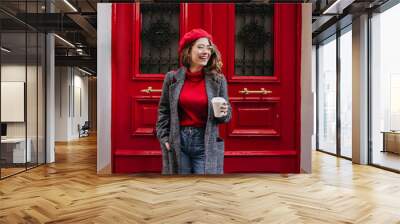 Glad white female model in long gray jacket looking away with smile standing near red door. Outdoor photo of beautiful caucasian woman in sweater drinks coffee in front of cafe and laughing. Wall mural