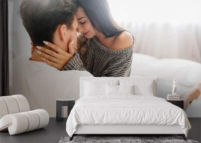 Glamorous tanned girl with white manicure resting with husband in sunday morning. Fascinating european woman holding boyfriend's face with tenderness, sitting on bed. Wall mural