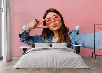 Glamorous young woman taking selfie on pink background. Appealing brunette girl showing peace sign. Wall mural