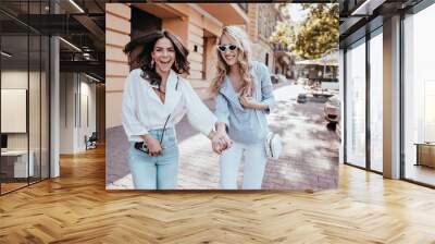 Gorgeous brunette girl holding hands with friend while walking around city. Outdoor portrait of two laughing sisters spending leisure time together. Wall mural