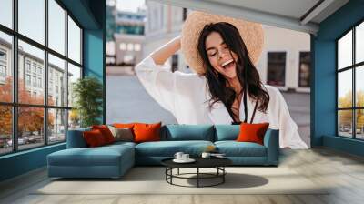 Gorgeous brunette woman wears white blouse smiling on street background. Beautiful laughing girl in straw hat spending day in city. Wall mural