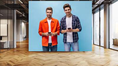 Handsome young charming men in orange jacket and checkered shirt smile sincerely and hold phone on blue background. Wall mural