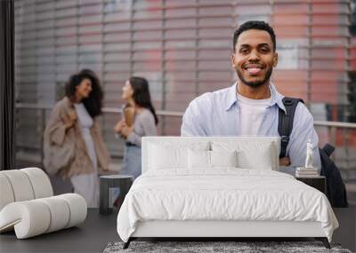 Happy african young guy posing looking at camera on background of students at university. Man wearing shirt is standing outdoors and smiling. Lifestyle concept Wall mural