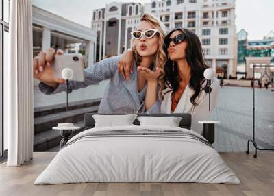 Happy blonde girl in white sunglasses making selfie with kissing face expression. Glamorous female friends expressing love for photo. Wall mural