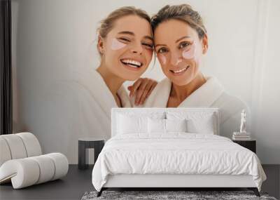 Happy caucasian mother with daughter wearing hydrogel patches smiling looking at camera on white background. Blondes wear robes to take care of their skin. Cosmetic natural trend. Wall mural