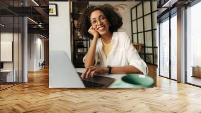 Happy young african woman looking at camera sitting at laptop home. Brunette with curly hair wears white shirt. Mood, lifestyle, concept. Wall mural