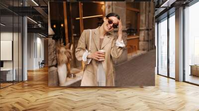 Happy young caucasian girl walking with cup of coffee in city on sunny day. Brunette wears stylish jacket, sunglasses and bag. Sincere emotions lifestyle concept Wall mural