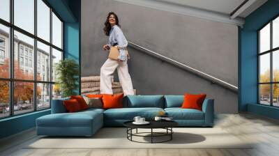 In full growth, young caucasian woman with documents in hands climbs stairs going to work. Brunette wears blue shirt, white trousers and sweatshirt. Business lady concept Wall mural