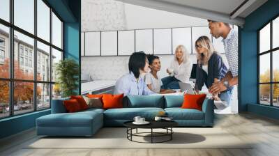 Indoor portrait of asian guy and african girl sitting at the desk in university and talking with friends. Young managers arguing about action play during meeting. Wall mural