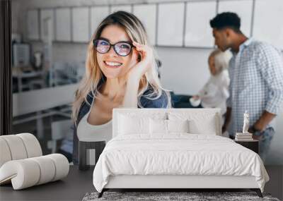 Indoor portrait of excited student girl in glasses and gray jacket. Attractive female employee posing in office and laughing with colleagues on background. Wall mural