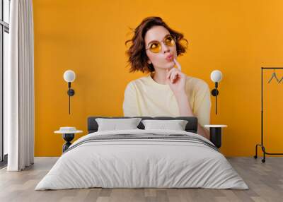 Indoor portrait of pensive brunette girl in light-yellow t-shirt. Glad short-haired woman in sunglasses looking up and thinking about something. Wall mural