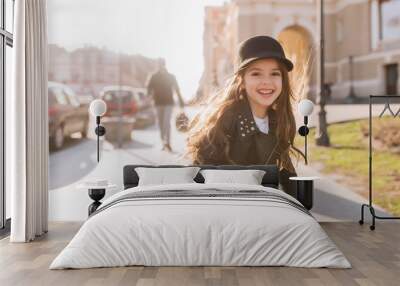 Inspired cute child with long dark hair playing on the street in good mood, waiting for friends and smiling. Portrait of pretty little girl in leather jacket posing on blur background in morning. Wall mural