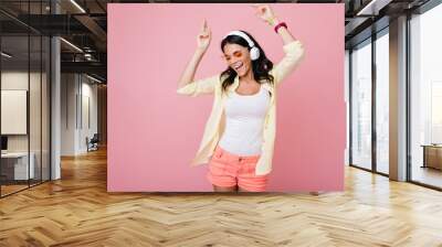 Inspired slim brunette girl in sunglasses funny dancing on pink background and waving hands. Laughing dark-haired young woman in yellow shirt enjoying music in headphones with eyes closed. Wall mural