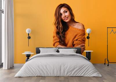 Interested long-haired lady in orange sweater looking to camera. Debonair european female model standing on yellow background with arms crossed. Wall mural