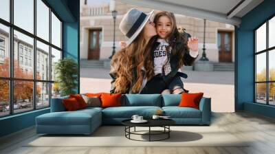 Joyful brunette girl with lovely face expression in stylish jeans with holes sitting on mom's knee and laughing. Beautiful woman wearing elegant hat kissing daughter in cheek in the middle of street. Wall mural