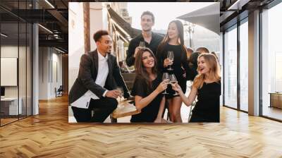 Joyful brunette man in black shirt looks away, holding glass of wine at party. Outdoor portrait of african guy in trendy jacket celebrating birthday with charming girls in cafe. Wall mural