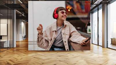 Joyful teen girl with brunette hair listening to music outside. Short-haired lady in red headphones, pink sunglasses and light outfit smiles in cafe.. Wall mural