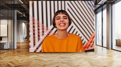 Joyful woman with brunette hair smiling widely on striped background. Trendy short-haired lady in orange sweatshirt posing outdoors.. Wall mural