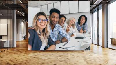 Laughing african student in blue shirt sitting with laptop between european university friends. Beautiful blonde female manager making selfie during conference with colleagues. Wall mural