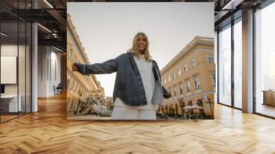 Mischievous young caucasian woman wears white sweatshirt, denim jacket in summer or autumn. Blonde girl stands in front of camera outdoors. Trendy background with copy space. Wall mural