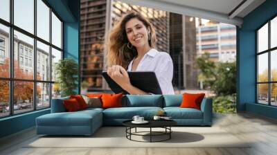 Nice young caucasian business woman working with tablet computer on city street. Blonde looks at camera, wears white shirt. Concept of education, technology Wall mural