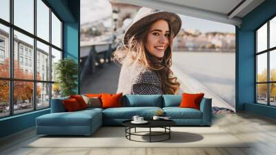 Outdoor photo of romantic european woman with curly hairstyle spending time outdoor, exploring european city. Graceful young lady in gray coat and hat enjoying views on embankment. Wall mural