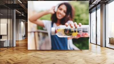 Outdoor portrait of smiling black girl in white shirt with bottle in her hand on foreground. Blur photo of attractive young woman with curly hair posing with water on tree background. Wall mural