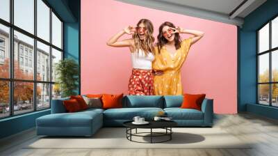 Photo of two young caucasian women in bright clothes with polka dots standing on pink background. Blonde and brunette in sunglasses are smiling broadly with their teeth and showing sign of peace. Wall mural