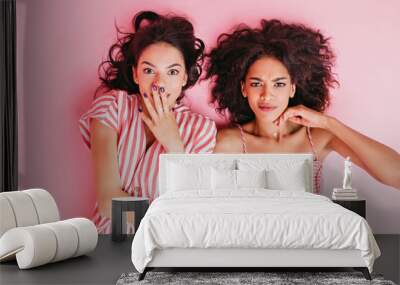 Photo on isolated pink background in studio. Two girls with beautiful tan and dark curly hair lie on their backs and pose in front of camera with pathetic look Wall mural