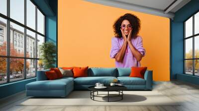 Pleased black girl in trendy sunglasses having fun during indoor photoshoot. Refined stylish african woman playfully smiling on yellow background. Wall mural