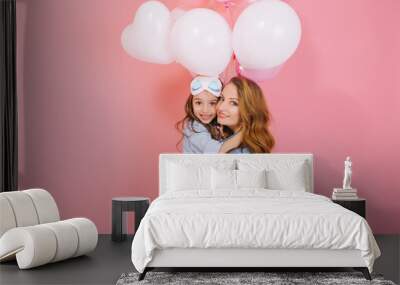 Portrait of cute long-haired little birthday girl with white balloons embracing her young curly mom after event. Charming mother posing with pretty daughter at party isolated on pink background Wall mural