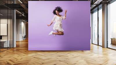 Portrait of funny african girl in white attire jumping on purple background. Studio shot of blithesome brunette young woman expressing positive emotions. Wall mural