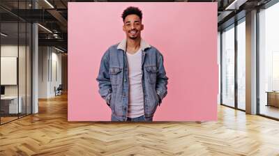 Portrait of smiling man in denim jacket looks into camera. Handsome brunette guy in jeans and white tee widely smiles on pink background Wall mural