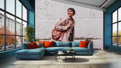 Positive hipster with short hair smiling outdoors. Brunette girl with brown backpack in beige jacket posing on backdrop of white wall.. Wall mural