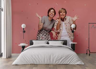 Positive two ladies with short trendy hairstyle in light stylish outfit smiling and posing with confetti on pink background.. Wall mural