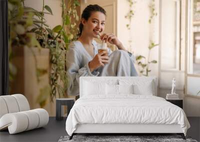 Positive young caucasian woman looks at camera, drinks delicious cocktail sitting indoors. Brunette wears long blue sundress in summer. Mood, lifestyle, concept. Wall mural