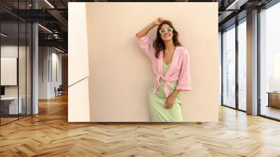 Positive young caucasian woman smiling with teeth posing at camera near building wall. Model wears shirt, sundress and sunglasses. Concept of stylish vacation. Wall mural