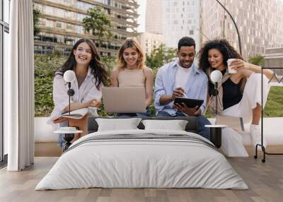 Positive young diverse classmates use laptop and tablet for study sitting outside. Guy, girls with good mood wear casual clothes. Concept of journalism, learning. Wall mural