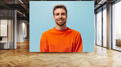 Positive young guy with red beard in bright orange sweatshirt looking into camera and smiling on isolated blue background.. Wall mural