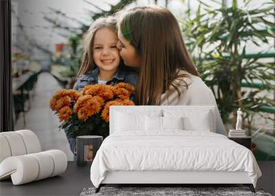 Pretty little girl smiling while her mom kissing her. Photo of caucasian family spending time in orangery. Wall mural