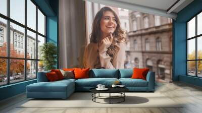 Pretty white woman playfully touching chin with finger while looking at city. Indoor photo of laughing romantic girl with cup of coffee standing near window. Wall mural