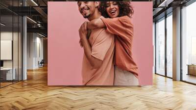 profile photo of young woman hugging from behind tall guy indoors in pink. happy brunettes with curly hair are dressed in light summer clothes. Wall mural