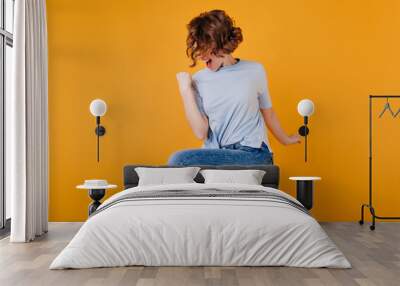 Refined short-haired lady wears dark-blue jeans jumping in studio. Attractive girl with wavy hairstyle dancing on yellow background. Wall mural