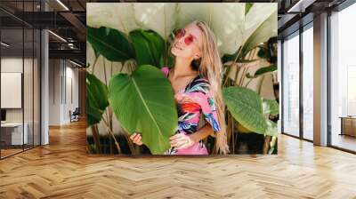 Romantic girl in bright stylish attire enjoying photoshoot on nature background. Photo of adorable blonde woman with happy face expression touching green leaf. Wall mural