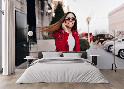 Romantic pretty woman posing with hair waving while walking outdoor. Photo of positive european girl in casual red attire talking on phone. Wall mural