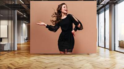 Self-confident lady in short black dress laughs and plays her hair on beige background Wall mural