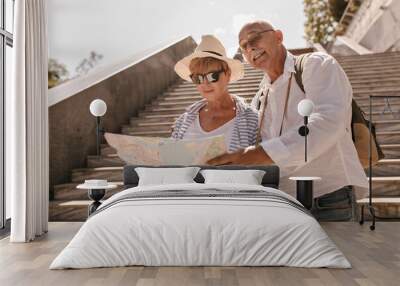 Short haired lady in hat, sunglasses and striped blouse holding map and posing with man in mustache in white shirt on background of stairs.. Wall mural