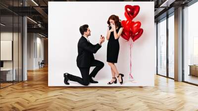 Shy girl in black dress posing in valentine's day. Brunette cheerful man standing on one knee while making proposal. Wall mural